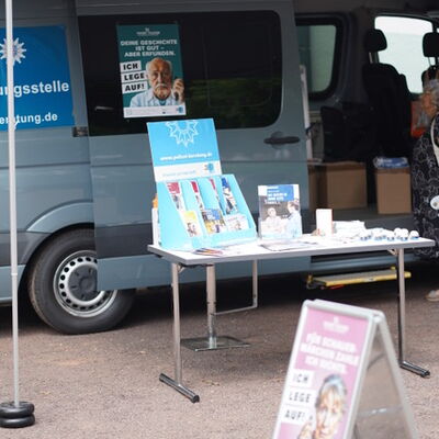Ein Infostand mit ausgelegten Broschüren und Handzetteln vor einem Transporter. Im Vordergrund ein Plakataufsteller mit einer älteren Person und dem Slogan: 'Ich lege auf'