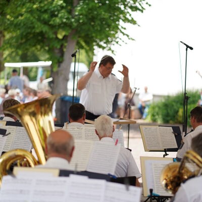 Das Polizei Orchester mit Blechblasinstrumenten von hinten fotografiert, während der Dirigent im weißen Hemd im Hintergrund das Ensemble unter Bäumen im Freien leitet.